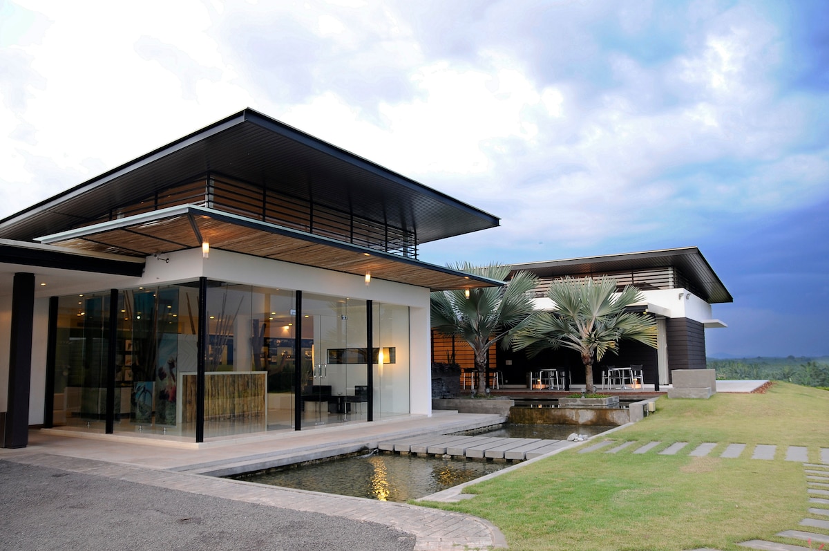 The event space features modern architecture with large glass panels that invite natural light. A tranquil pond is located near the entrance, surrounded by lush green grass. Palm trees provide shade, while outdoor seating areas offer a relaxed setting for gatherings.