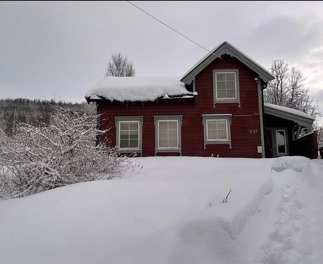 The red house is partially covered in snow, with a sloped roof and a front porch. Nearby, snow-covered bushes contribute to the winter landscape. Three windows decorate the front, allowing natural light to fill the interior. The surrounding area features a tranquil blanket of snow.