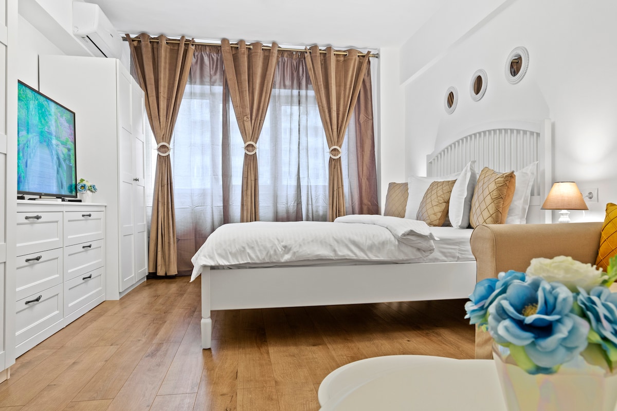 A bright bedroom features a spacious white bed with neatly arranged pillows. Natural light comes through large windows covered by elegant, flowing curtains. A cozy sofa is positioned beside the bed, and a dresser with a TV can be seen against the wall, complemented by decorative elements.