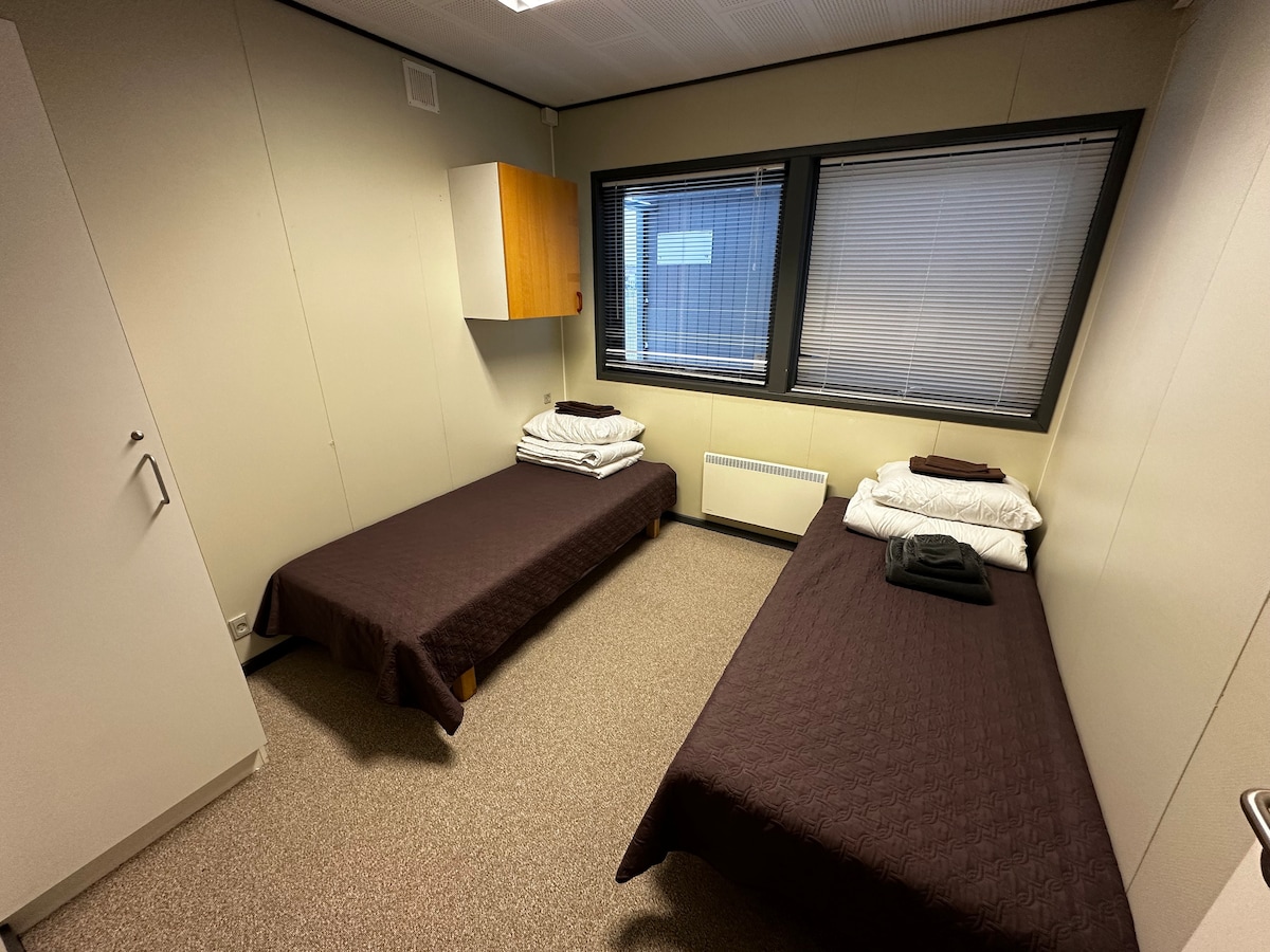 A room with two single beds, each topped with white pillows and dark brown blankets. A wall-mounted cabinet is positioned above one bed, and a window with blinds allows natural light to enter. A grey towel is neatly folded on one bed.