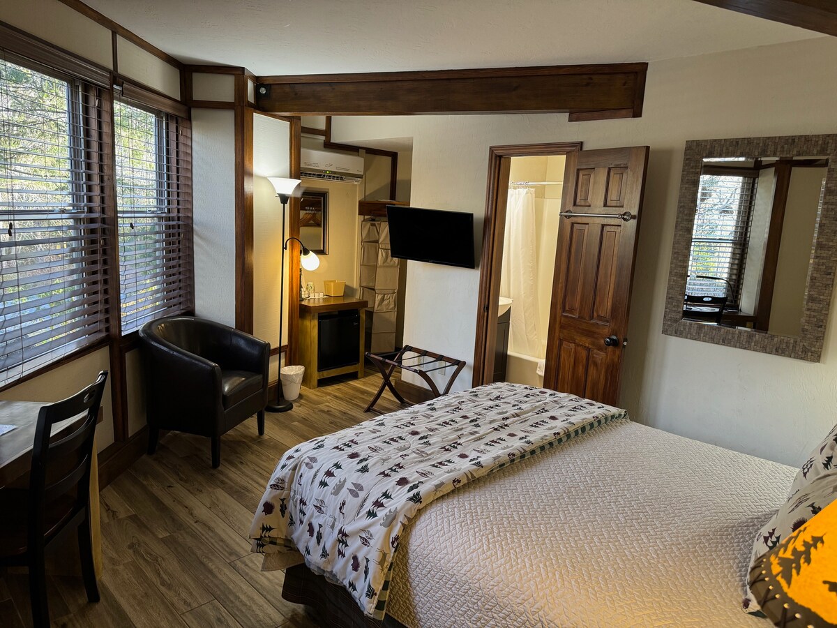 A spacious room features a queen bed with a patterned throw blanket. Two windows allow natural light to illuminate the area. A flat-screen TV is mounted on the wall opposite a small seating area with a chair and table. A private bathroom is visible in the background.