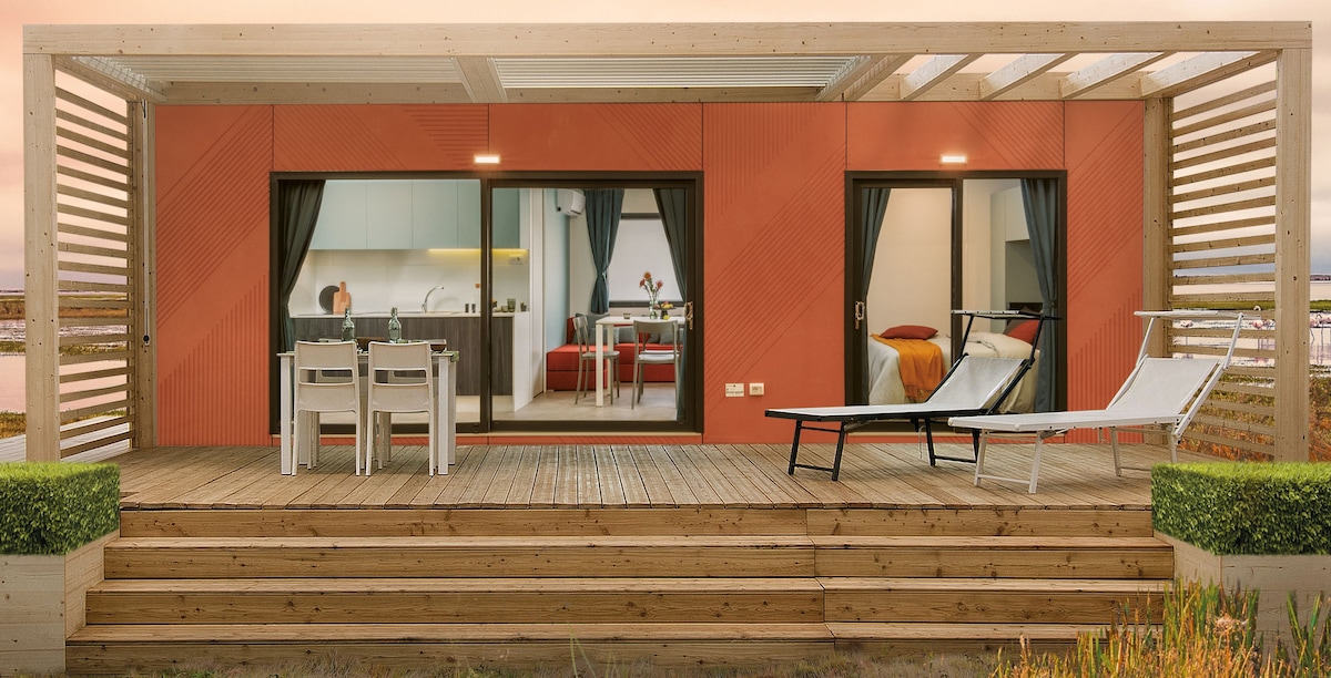 A spacious deck is showcased with wooden steps leading to an outdoor area which includes modern lounge chairs and a dining set for four. Large glass doors provide a view into the interior rooms, highlighting a minimalist kitchen and a cozy sleeping area.