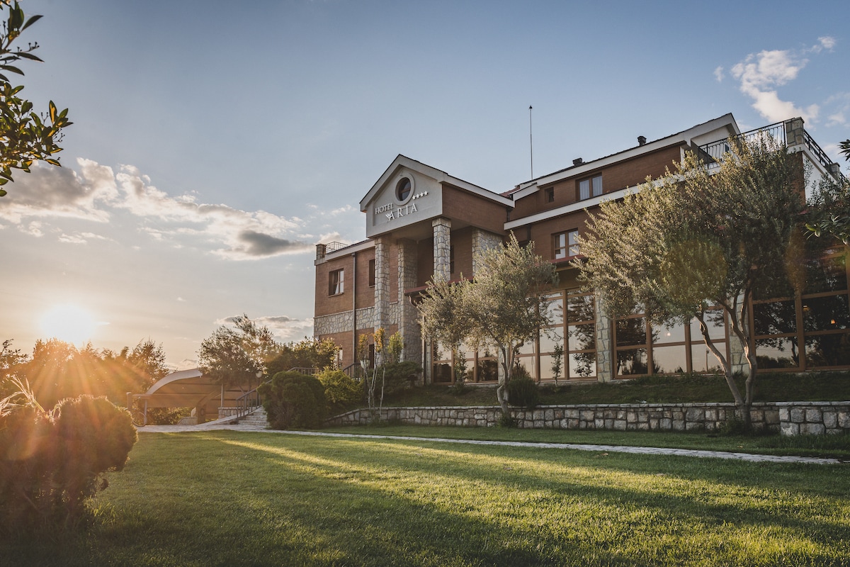 The exterior of Hotel Aria is displayed, featuring a modern building framed by well-maintained greenery. Large glass windows reflect the setting sun, while trees and landscaped lawns create a serene environment. The structure showcases a welcoming design, inviting guests to explore.