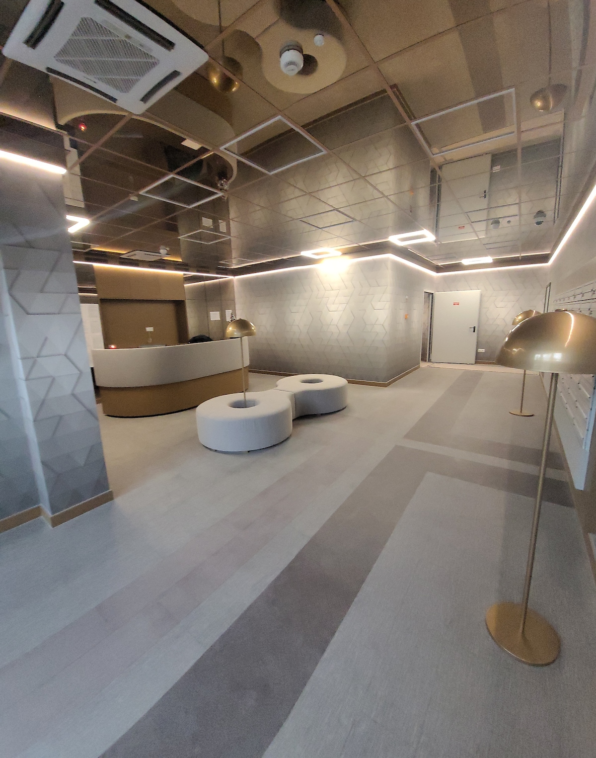A modern lobby area is highlighted by geometric wall designs and sleek ceiling lights. White circular seating is positioned around a stylish reception desk. Two golden floor lamps add a contemporary touch, illuminating the open space that connects to additional areas of the building.