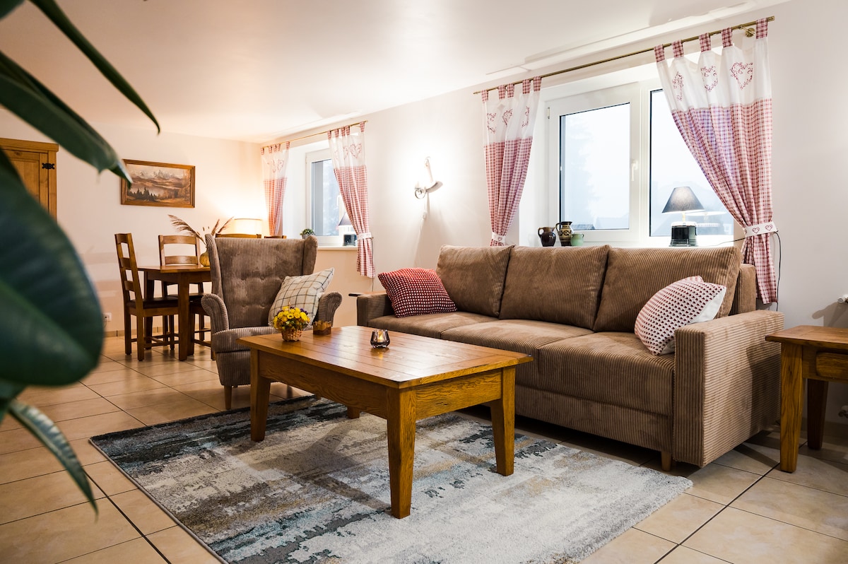 The inviting living area features a spacious sofa adorned with checked cushions, complemented by a rustic wooden coffee table. Light floods in through a window dressed with soft curtains, and a small dining table is positioned nearby, enhancing the cozy ambiance.
