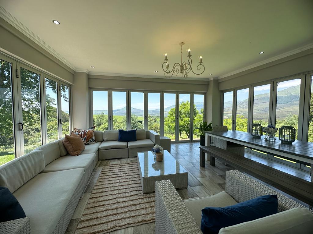 A spacious lounge area is designed with large windows showcasing expansive views of the surrounding landscape. A light-colored sectional sofa is complemented by a glass coffee table. A dining table is positioned nearby, inviting communal gatherings. Soft lighting enhances the calming atmosphere.