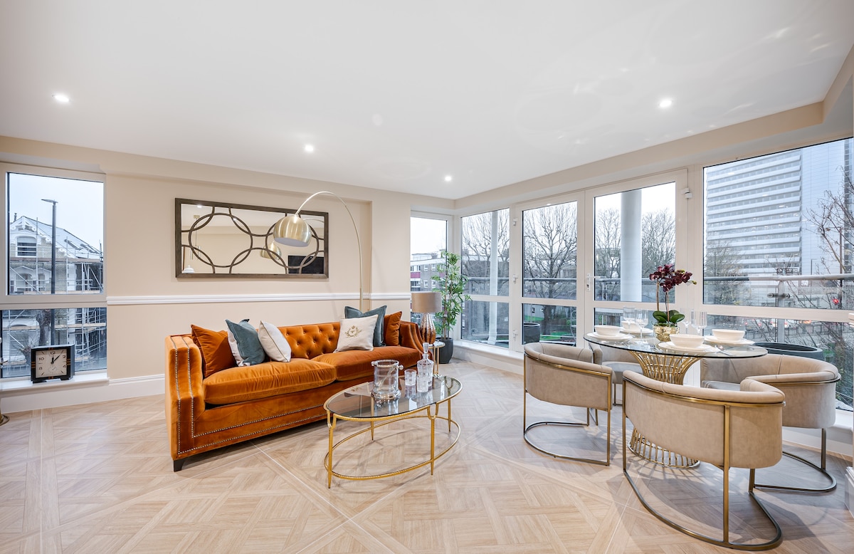 A spacious living area is presented with large windows allowing natural light to fill the room. A stylish orange sofa is paired with a glass coffee table and round dining table surrounded by four upholstered chairs. Decorative elements enhance the modern design.