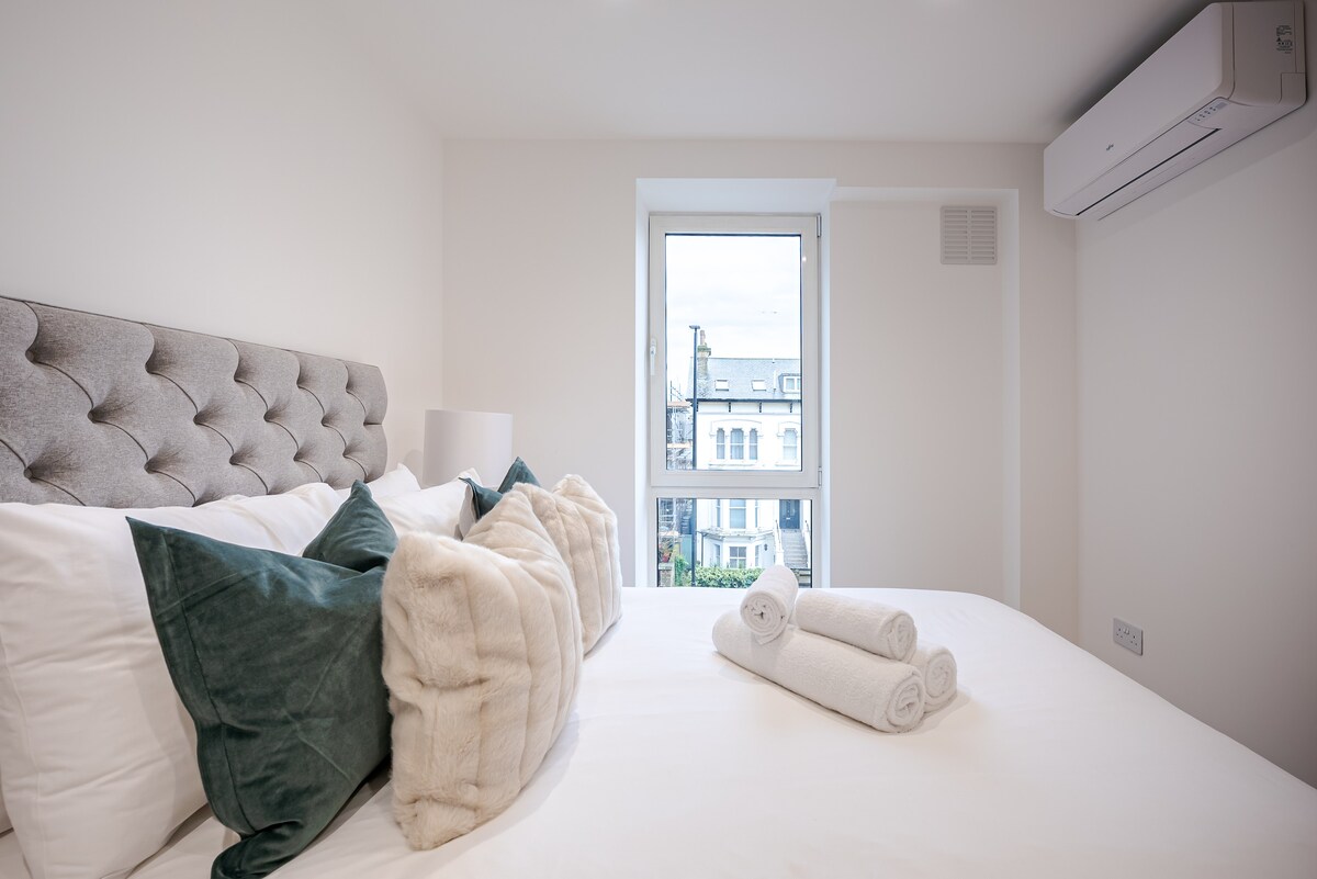 A spacious bedroom features a tufted headboard and soft linens, complemented by decorative pillows. Fresh towels are neatly arranged on the bed. A window offers a view of the outside, while a modern air conditioning unit is mounted on the wall.