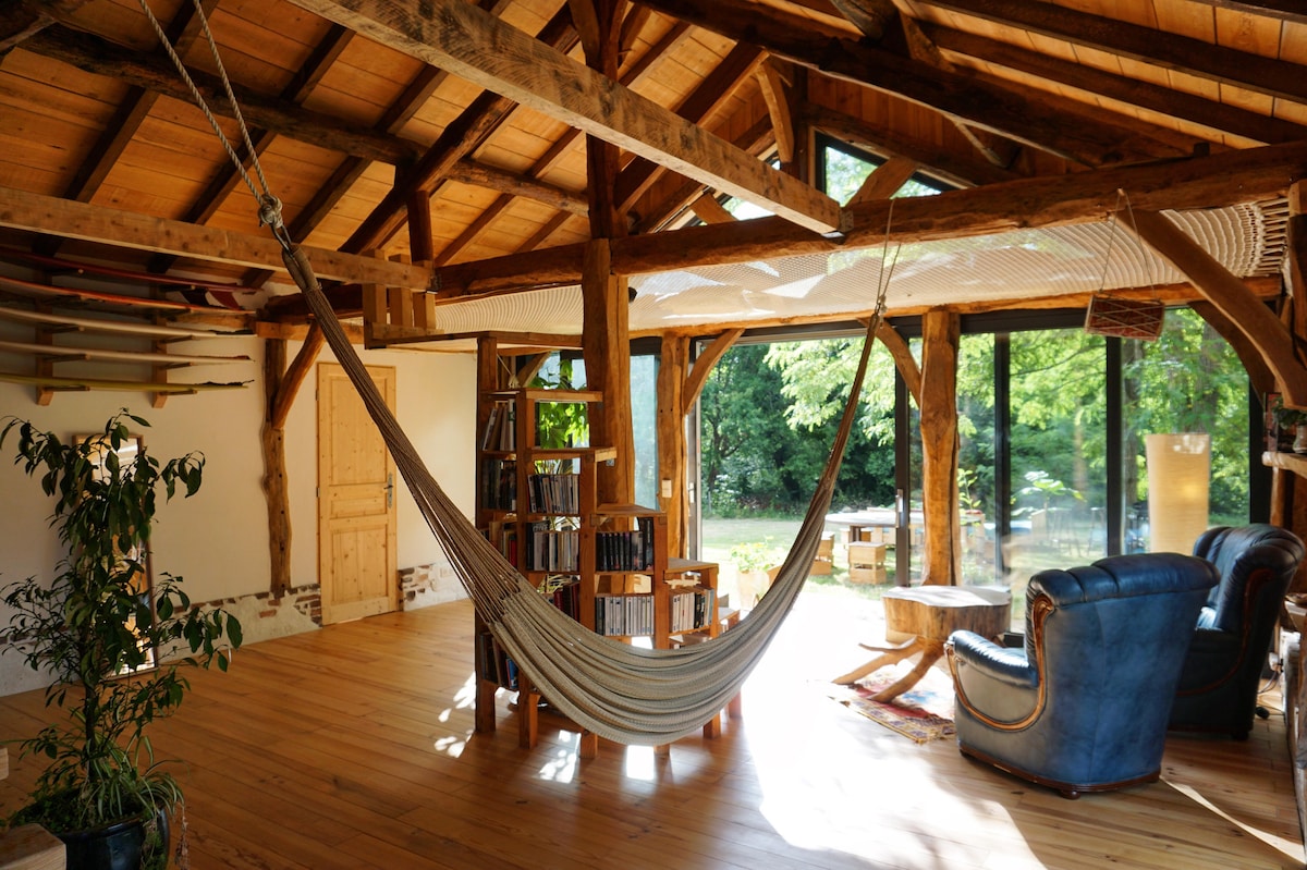 A spacious interior features exposed wooden beams and a hammock suspended from the ceiling. Natural light floods the room through large windows, highlighting a cozy seating area and a wooden bookshelf. Greenery outside can be seen through the glass, enhancing the serene environment.