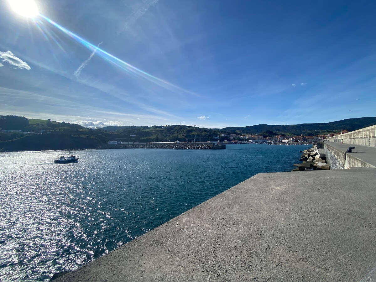 A wide view of the calm sea under a clear blue sky, showcasing gentle waves reflecting sunlight. The distant coastline features a charming town nestled among green hills. A boat is seen in the water, while the concrete pier extends into the sea on the right.