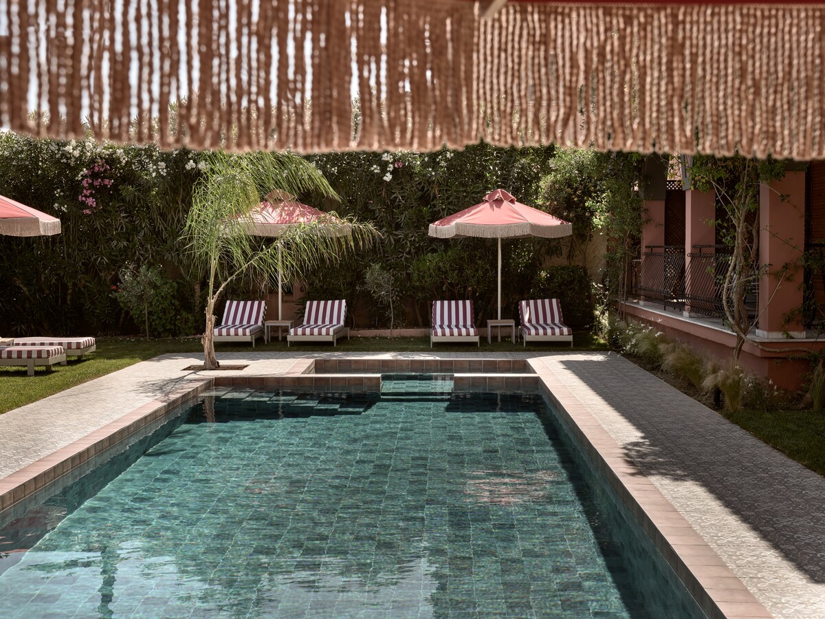 A tranquil swimming pool is surrounded by manicured greenery, featuring a surrounding deck with striped lounge chairs and umbrellas. The pool is partially shaded, reflecting the serene atmosphere of the outdoor space, which is enhanced by blooming flora in the background.
