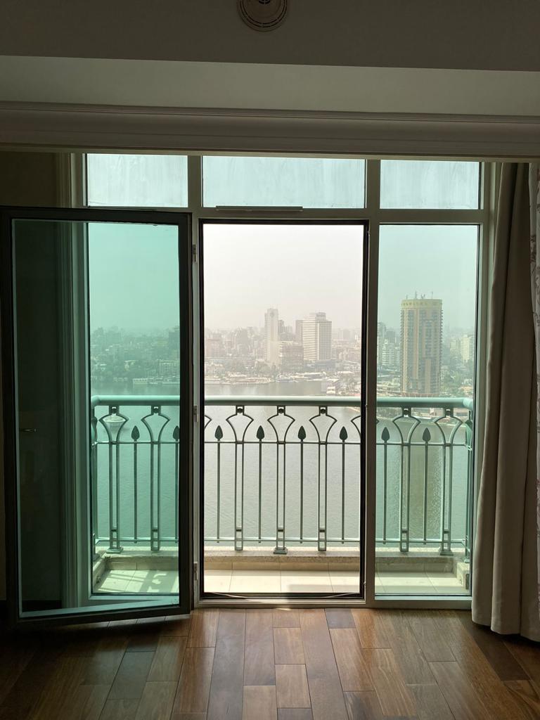 Floor-to-ceiling glass doors open onto a private balcony, revealing a panoramic view of the Nile and the Cairo skyline. The space features warm wooden floors and muted curtains that drape gracefully beside the doors, inviting natural light into the room.