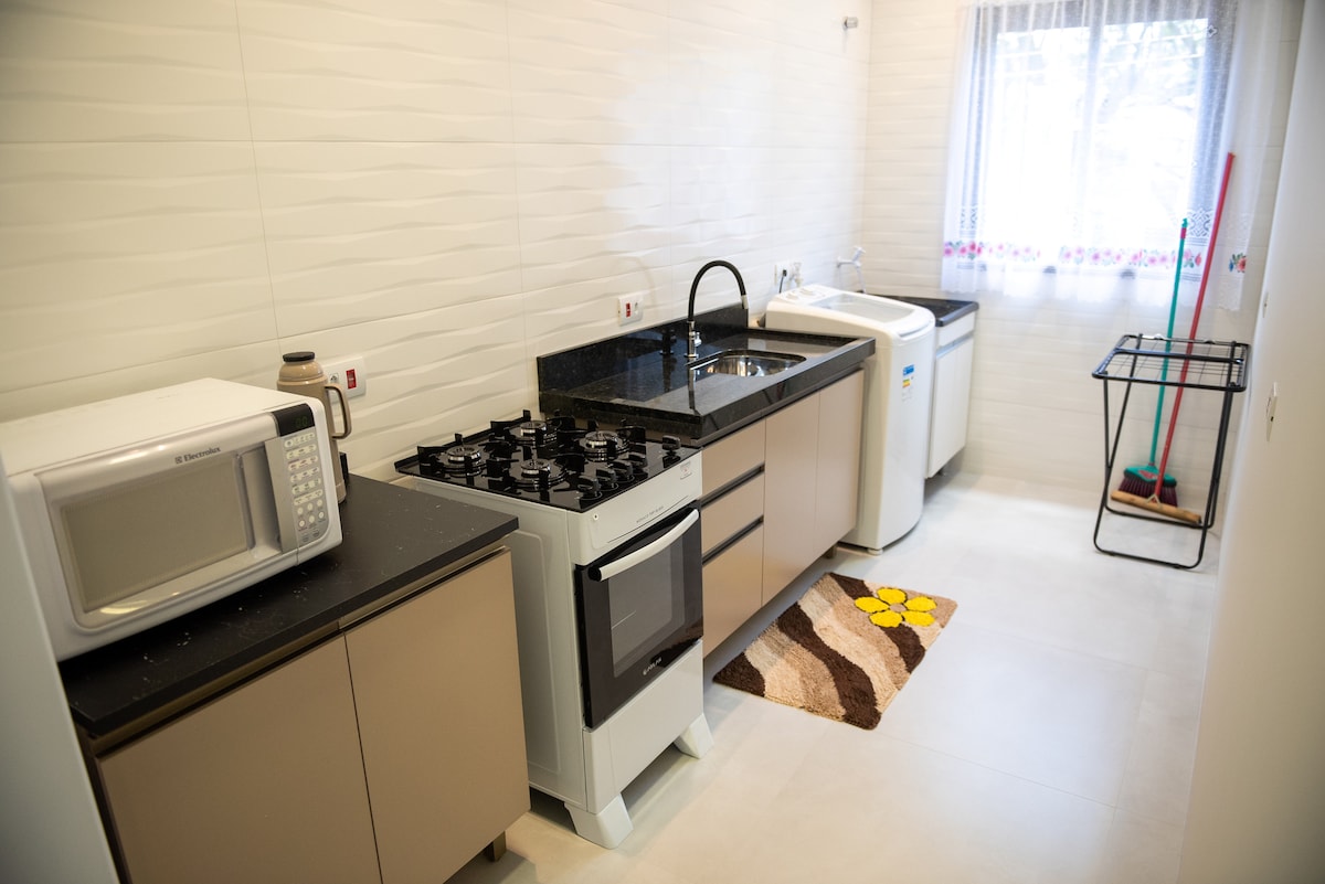 A modern kitchen features a black granite countertop with a stove and microwave present. A washing machine is located at the end of the counter, complemented by a small rug with a floral design. Natural light enters through a window adorned with a simple curtain.