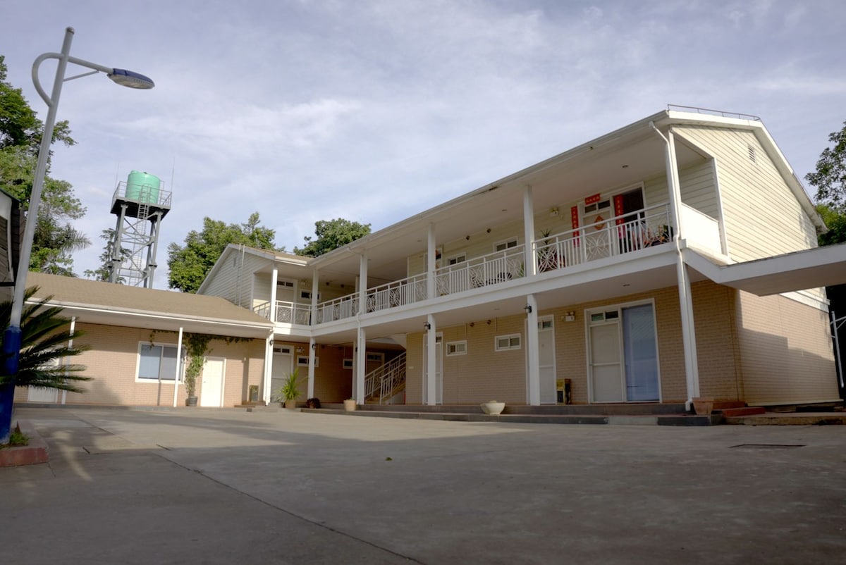 The exterior view showcases a two-story building with a classic design, featuring balconies along the upper level. A spacious paved area is visible in front, bordered by greenery. A water tower stands in the background, contributing to the practical setup of the property.