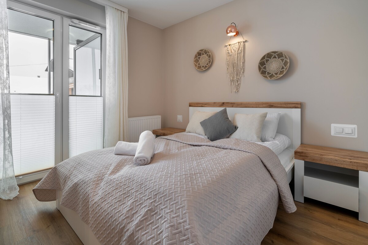 A serene bedroom is presented, featuring a spacious bed adorned with a textured blanket and pillows. Light-colored walls are accentuated by decorative wall art. Natural light enters through large glass doors that lead to a small balcony, enhancing the room's welcoming ambiance.