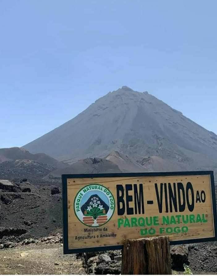 A large sign reading 'Bem-Vindo ao Parque Natural do Fogo' is positioned in the foreground. In the background, a majestic volcano rises against a clear sky, showcasing its rugged, volcanic slopes and distinct crater.
