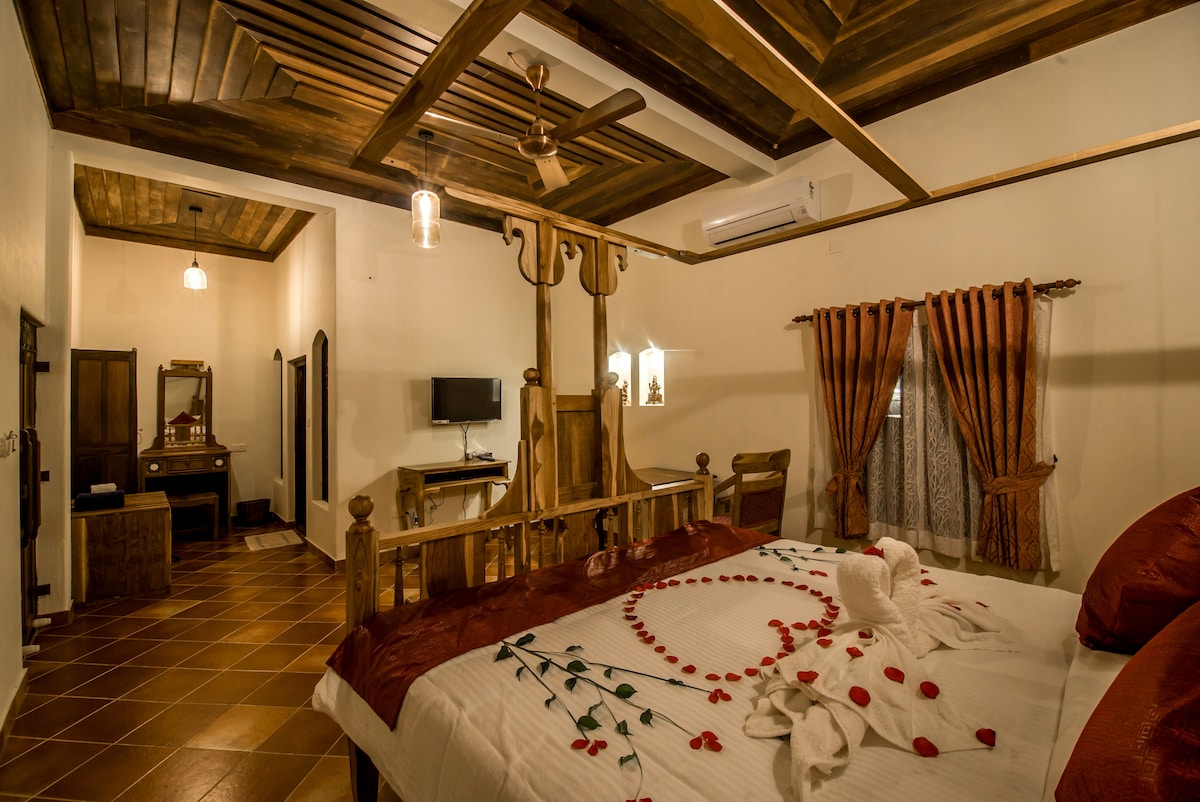 A spacious room features a large wooden bed adorned with flower petals, complemented by warm-colored bedding. An intricately designed wooden ceiling catches the eye, while a charming dresser and a small television are visible in the background. Soft curtains frame a window, adding to the inviting ambiance.