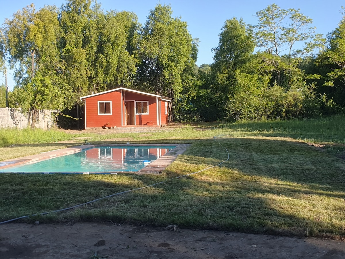 A serene outdoor space features a rectangular pool surrounded by freshly cut grass. In the background, a red cottage with white trim is framed by lush green trees, creating a tranquil setting ideal for relaxation.