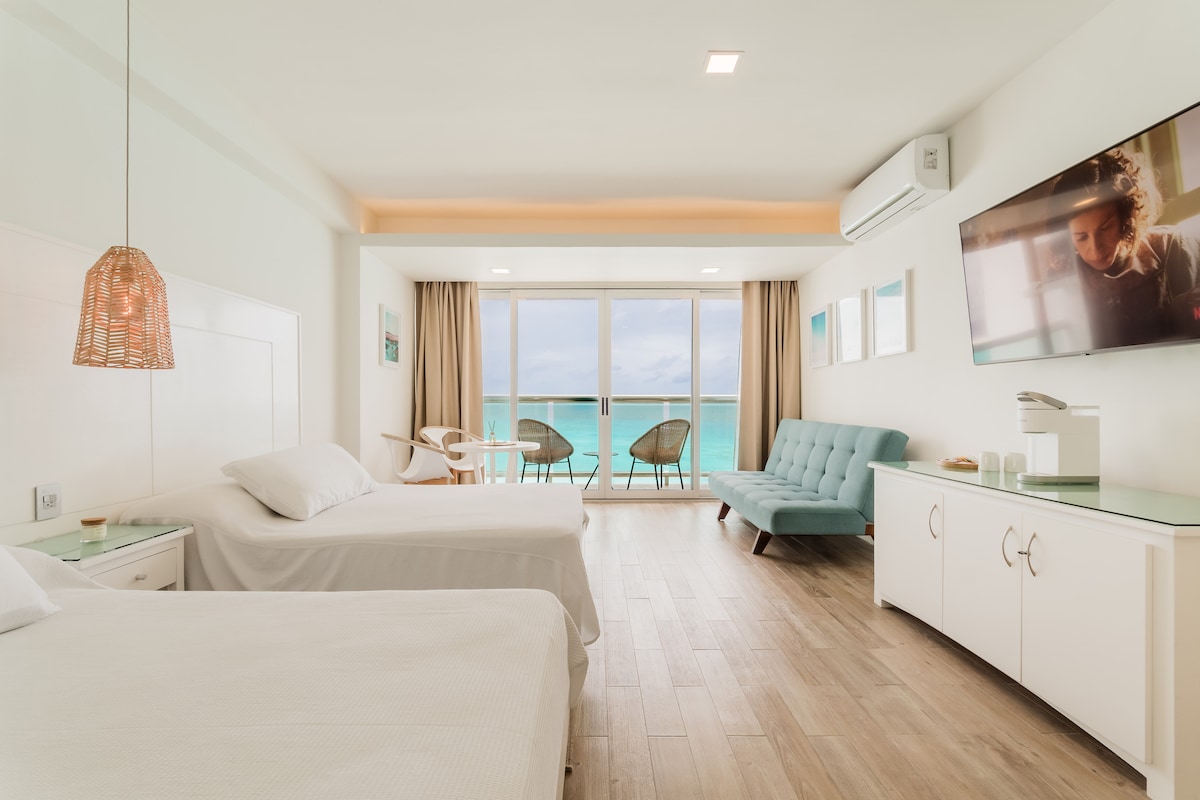 A bright and airy studio features two full beds and a comfortable sofa bed. A large balcony offers ocean views, while a flat-screen TV is mounted on the wall. Light wood flooring complements the simple decor, creating a relaxing atmosphere.