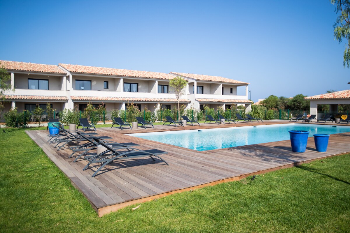 The image displays a serene outdoor area featuring a swimming pool surrounded by wooden decking. Lounge chairs are positioned neatly around the pool, with a clear blue sky overhead. The building is lined with lush greenery, enhancing the inviting atmosphere.