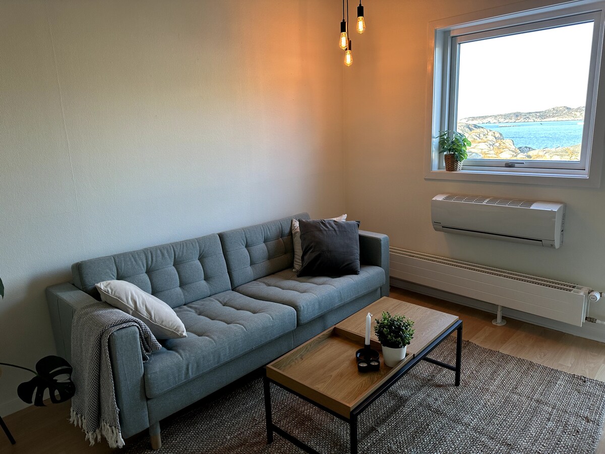 A light-filled living area features a modern sofa with tufted cushions and a decorative throw pillow. A wooden coffee table sits in front, complemented by a small potted plant. Large windows offer views of the ocean, enhancing the calm ambiance.