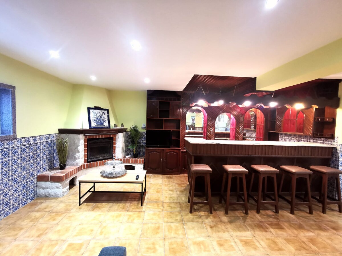 A spacious living area features a cozy fireplace and a wooden bar with seating for several guests. Various decorative elements, including traditional tiles, complement the warm-toned wall finishes. Soft lighting enhances the inviting setting, while a coffee table adds functionality to the space.