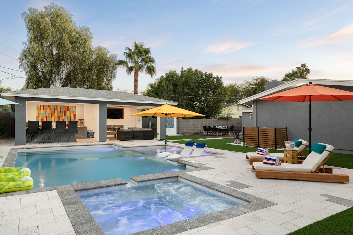 An inviting outdoor space features a heated pool and hot tub surrounded by lounge chairs. Colorful umbrellas provide shade, while a patio area with outdoor seating and a grill is visible in the background, enhancing the area for relaxation and entertainment.