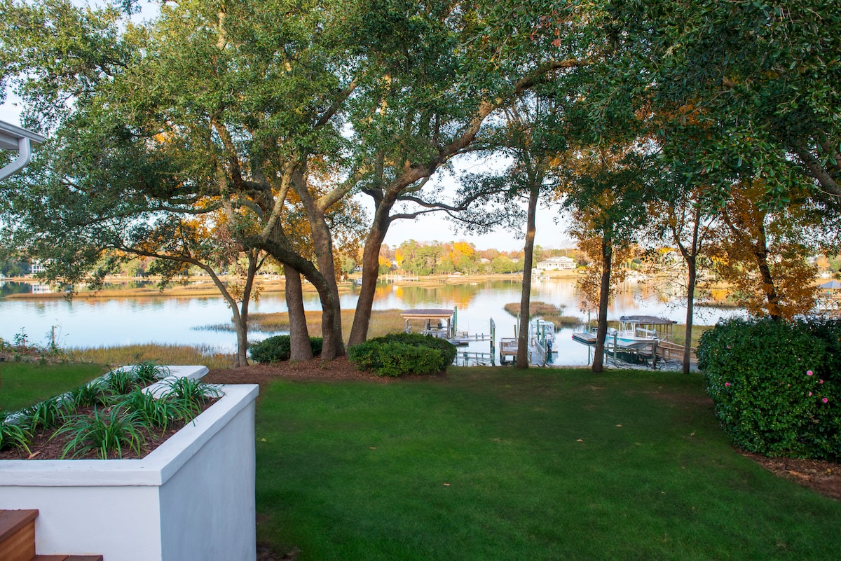 A serene view of the waterfront is framed by mature trees with colorful autumn foliage. A well-maintained lawn leads to a private dock, offering direct access to the water. Calm reflections of the trees and sky can be seen on the surface of the water.