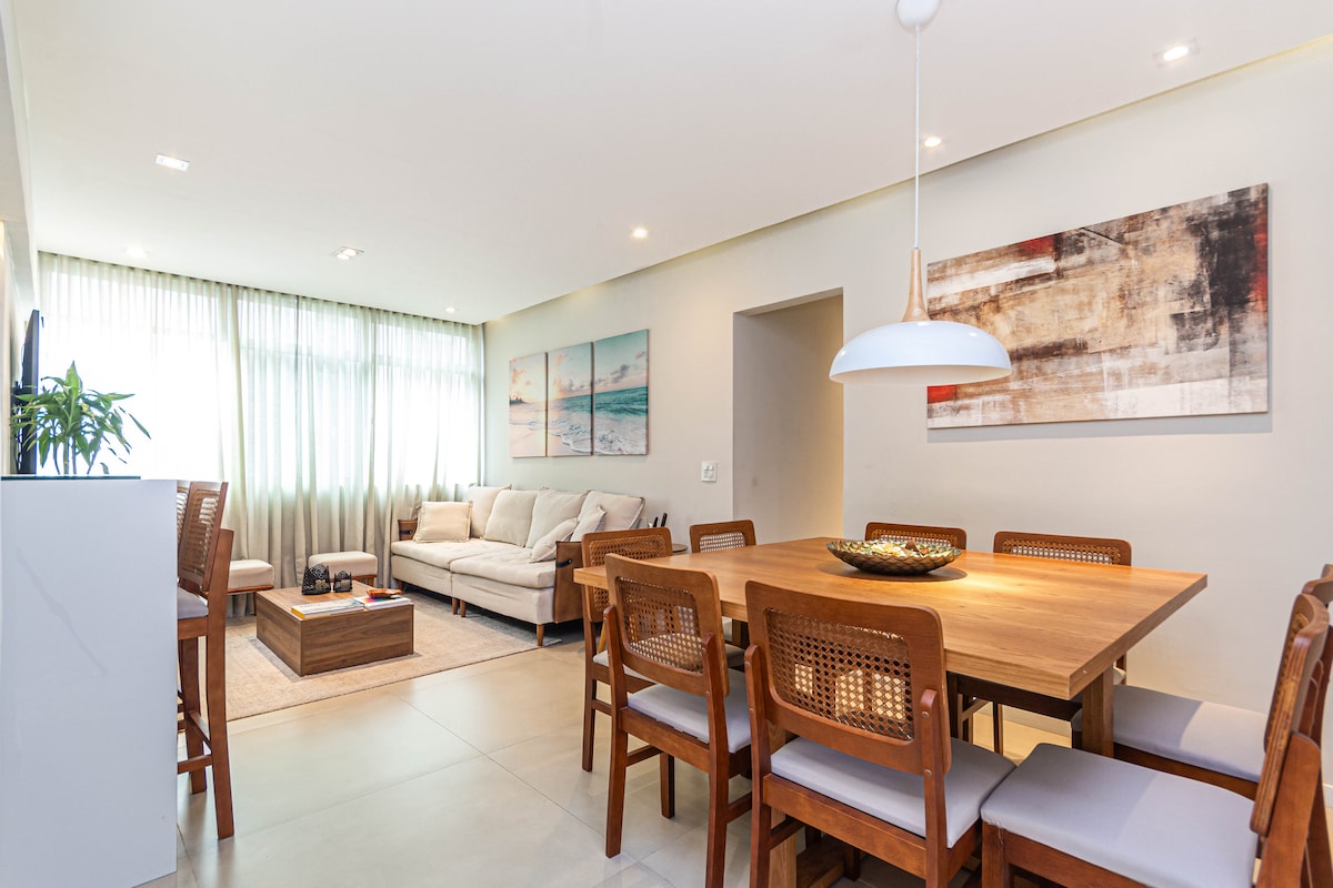 A spacious living area features a comfortable couch and a wooden dining table surrounded by chairs. Natural light filters through sheer curtains, illuminating the neutral-toned walls and decorative artworks that add a touch of character.