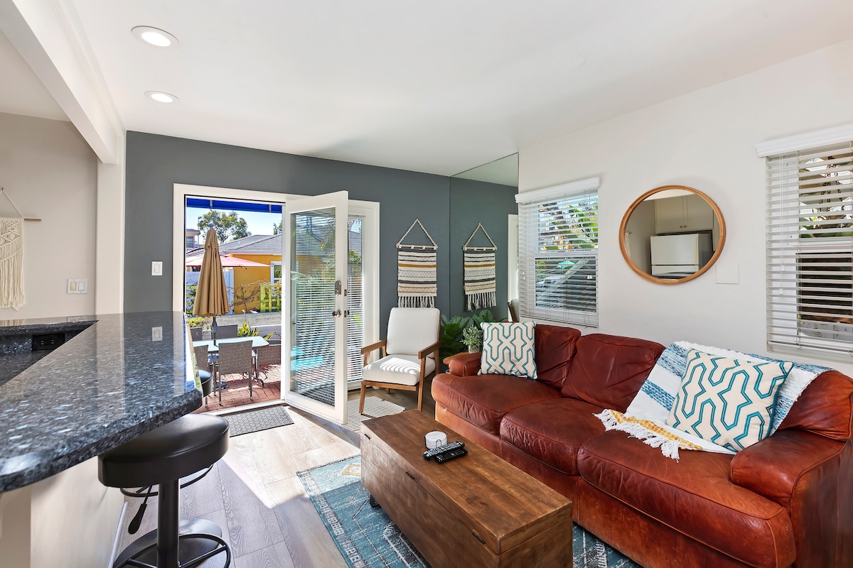 A cozy living area features a leather sofa and wooden coffee table, complemented by a patterned rug. Glass doors open to a patio, allowing natural light to fill the space. Decorative wall hangings add a touch of style, while a round mirror reflects the inviting ambiance.