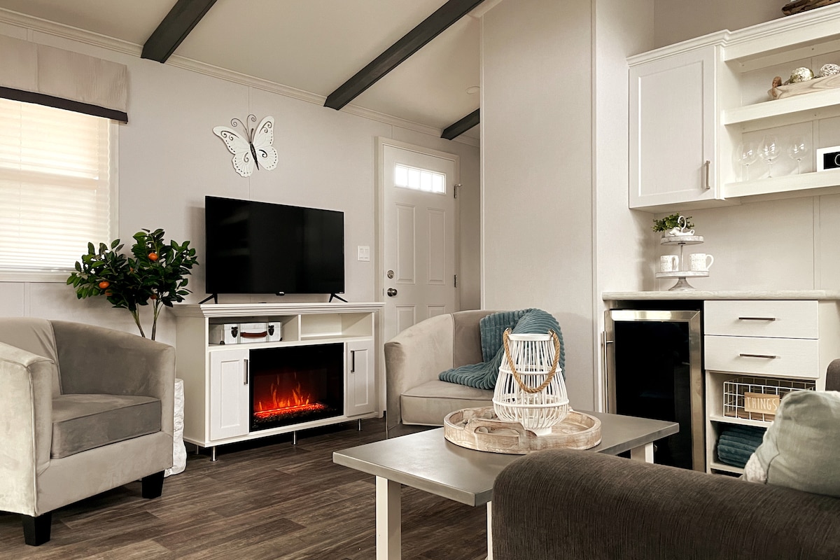 A cozy living space is presented with two plush armchairs and a comfortable couch. A modern electric fireplace adds warmth beneath a wall-mounted TV. A small table holds a decorative tray, while a compact fridge is positioned next to white cabinetry in the kitchen area.