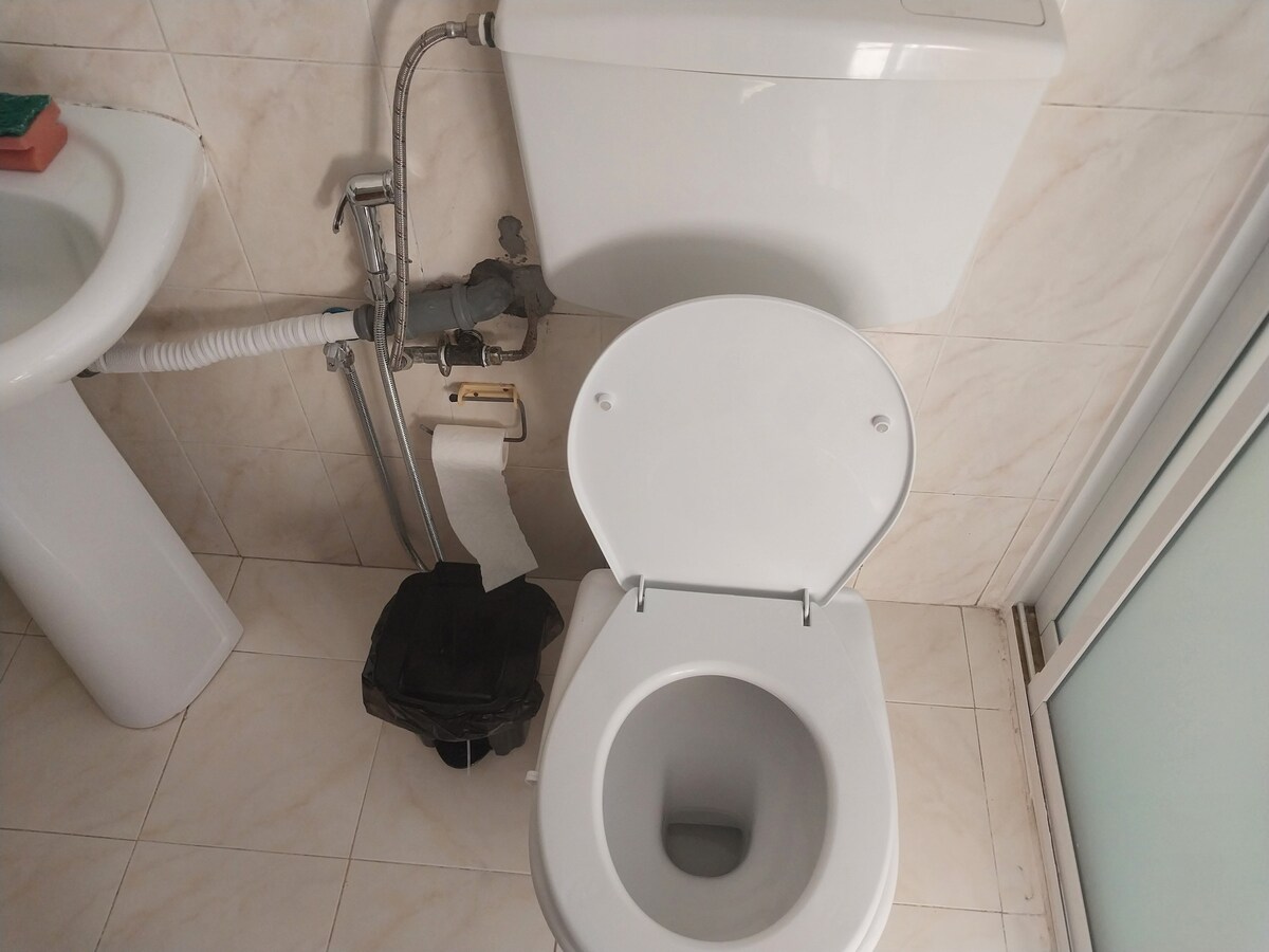 A clean and modern bathroom setting is shown, featuring a white toilet and a sink. Neutral tiles cover the floor and walls, providing a simple and functional appearance. A roll of toilet paper is neatly placed beside the toilet for convenience.