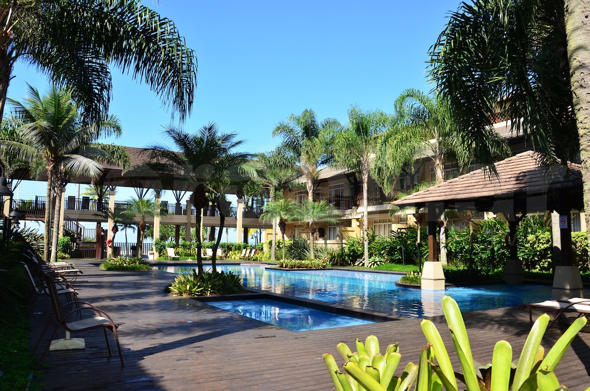 A serene pool area is surrounded by lush greenery, featuring various palm trees and vibrant plants. Sun loungers are positioned along the wooden deck beside the water, and spacious buildings are visible in the background, contributing to a relaxed atmosphere.