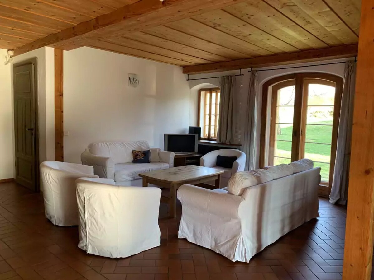 A communal area features a light-colored, comfortable seating arrangement consisting of four armchairs and a sofa, surrounding a rectangular wooden coffee table. A television setup is present, and large windows allow natural light to illuminate the space while providing views of the grassy area outside.