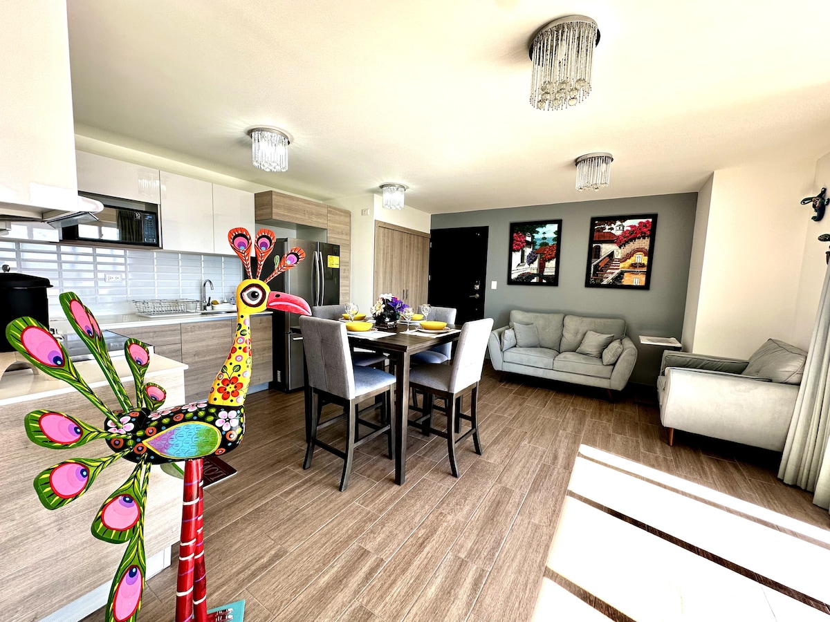 A modern living space is highlighted by a vibrant decorative sculpture. The area includes a dining table set for four and a comfortable seating arrangement. Artistic wall decor adds character, while natural light streams in through large windows, enhancing the contemporary design.