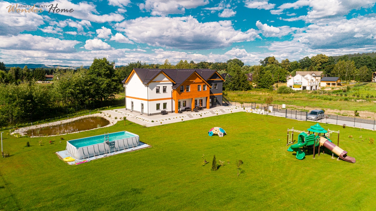 The image showcases a spacious outdoor area featuring a well-maintained lawn, a swimming pool, and a playground. Two distinct residential buildings are visible, with one featuring an orange façade. The sky is partly cloudy, contributing to a serene atmosphere.