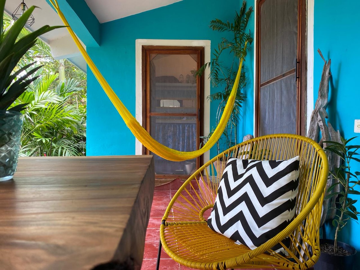 A vibrant outdoor area features a yellow hammock hanging beside a wooden table. A black and white striped cushion is placed on a wicker chair. Lush greenery surrounds the space, creating a natural and relaxing environment.