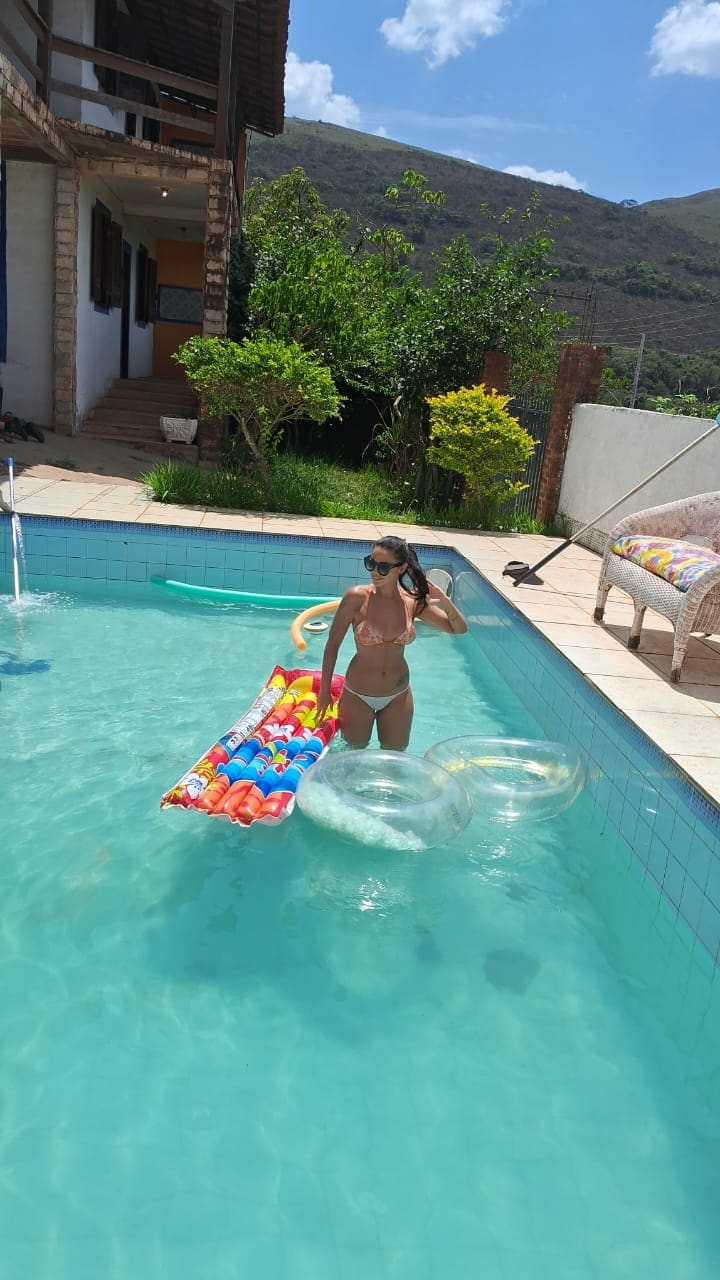A sparkling pool is surrounded by a patio with loungers. Colorful pool floats are seen in the water, while lush greenery provides a natural backdrop. A two-story house can be seen in the background, with stairs leading towards the entrance.
