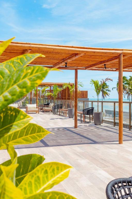 A rooftop area features wooden pergolas providing shade over modern seating arrangements. Lush greenery and palm trees line the space, with a clear view of the ocean in the background. The flooring consists of light-colored tiles, creating an open and airy atmosphere.