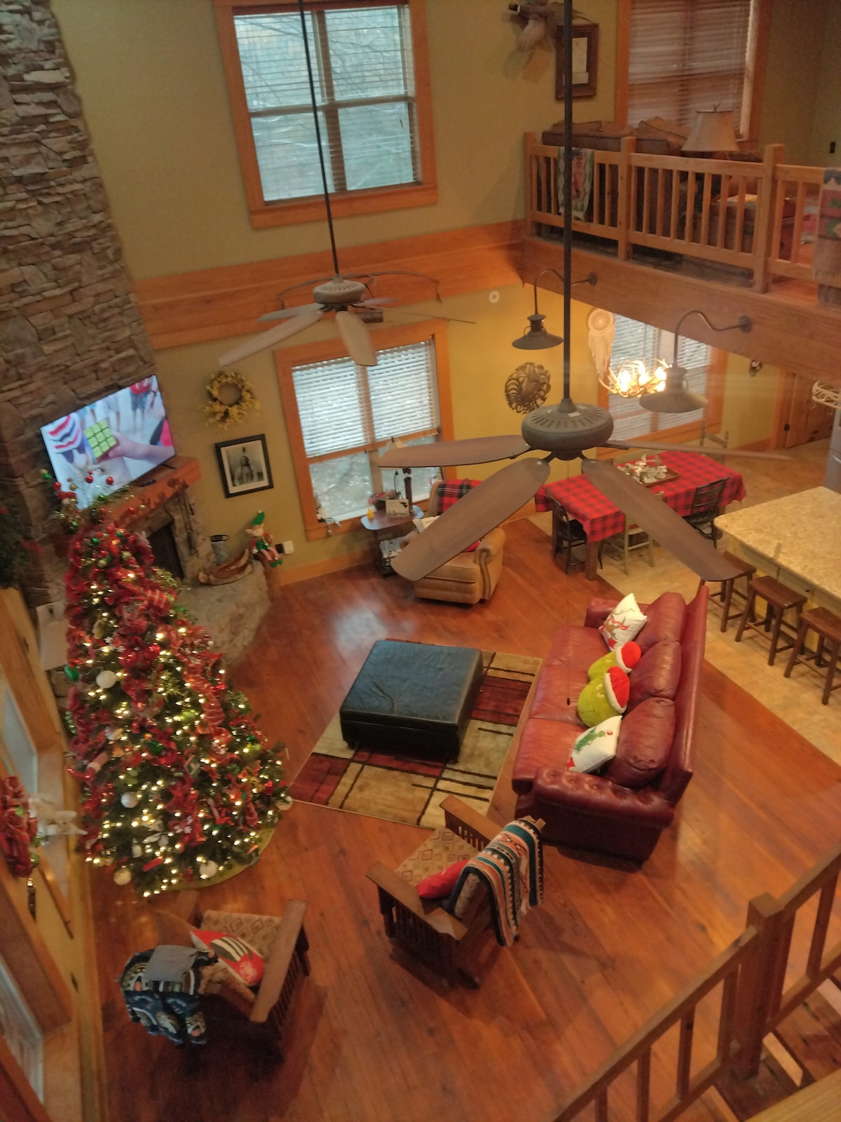 An open living space showcases a large sectional sofa adorned with festive cushions. A stone fireplace integrates with a mounted television. Nearby, a dining area features a long table, surrounded by several wooden chairs. A decorated Christmas tree stands majestically, contributing to the warm ambiance.