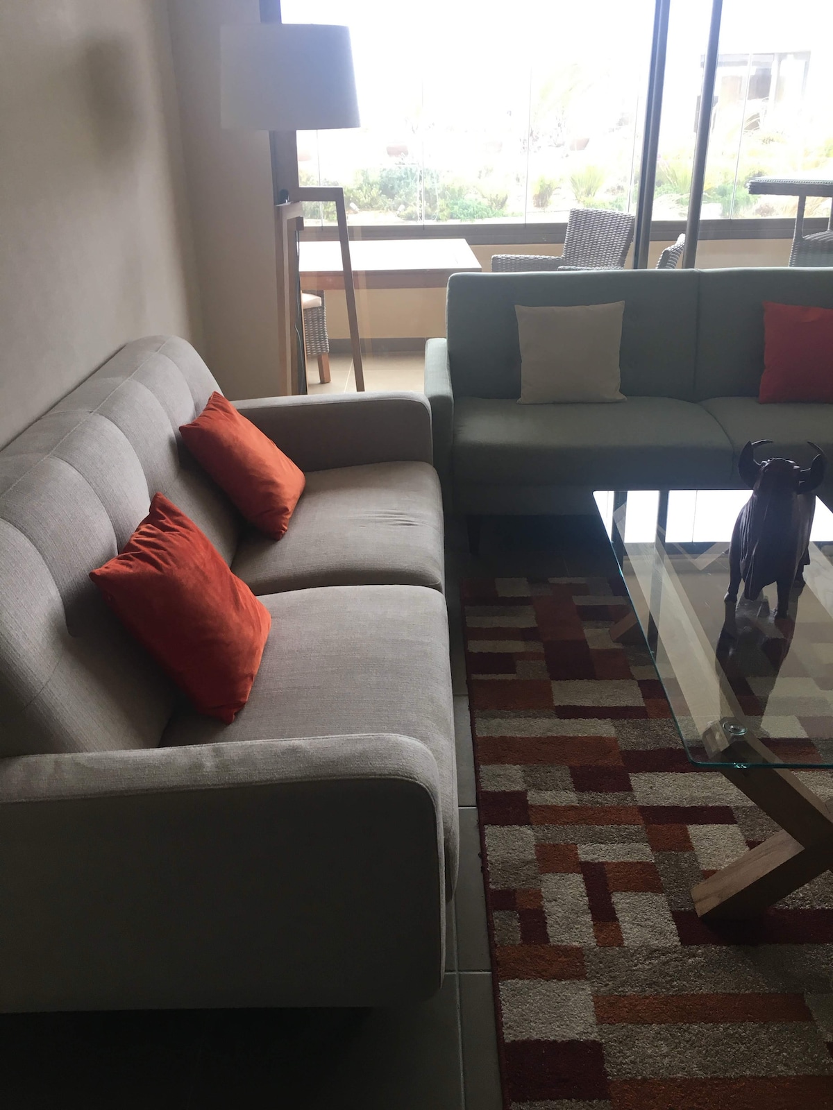 A comfortable living area features a light grey sofa adorned with orange cushions and a modern armchair. A glass coffee table stands in front, with subtle decorative elements placed atop it. Natural light enters through large windows, illuminating the space.