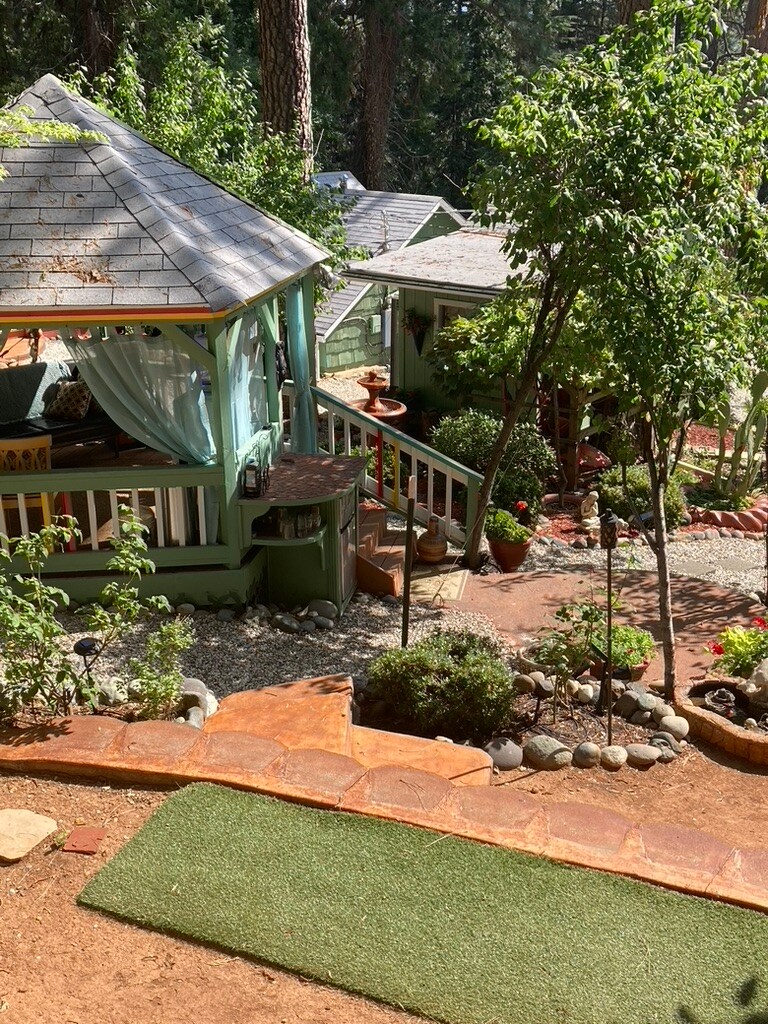 A scenic view showcases a charming gazebo surrounded by lush greenery and various potted plants. Pathways made of stone and gravel weave through the garden, leading to the cozy cabin. Sunlight filters through the trees, creating a serene and inviting outdoor space.
