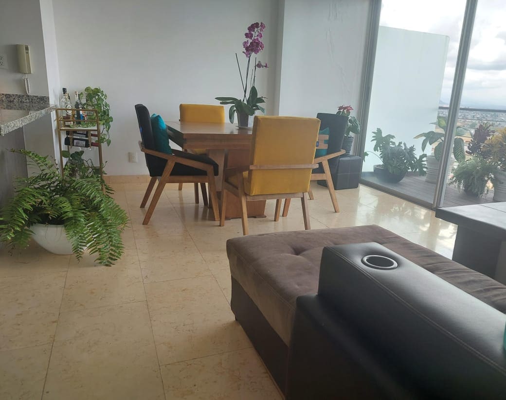 A bright dining area features a wooden table surrounded by six chairs in various colors. Potted plants add greenery, while large windows offer expansive views of the outside. A cozy lounge chair with built-in cup holder is positioned nearby, enhancing the inviting setting.