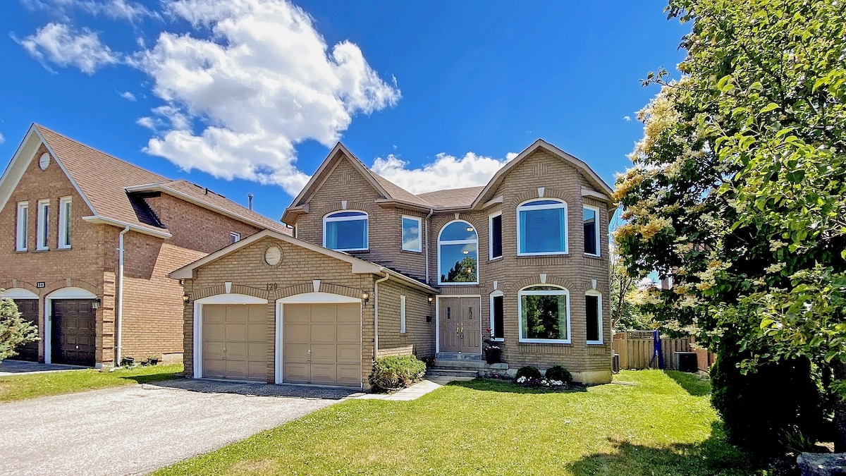 A two-story brick house stands in a tranquil neighborhood under a clear blue sky. The exterior features multiple large windows and a welcoming front entrance. A well-maintained lawn surrounds the house, with hedges and flowers adding greenery. The driveway accommodates two parking spaces.