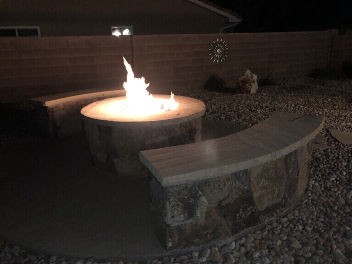 A circular stone fire pit is surrounded by a curved seating area, featuring smooth stone benches. The fire burns brightly in the center, providing a warm glow against the night sky. Pebbled ground surrounds the area, adding texture to the outdoor space.