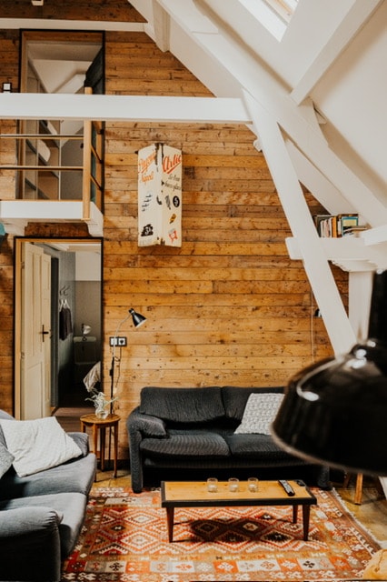 A cozy living area features wooden walls and a mix of comfortable seating. A decorative rug adds color to the floor, while a vintage-style hanging light fixture provides ambient illumination. A small table holds a few candles, and a door leads to another room.