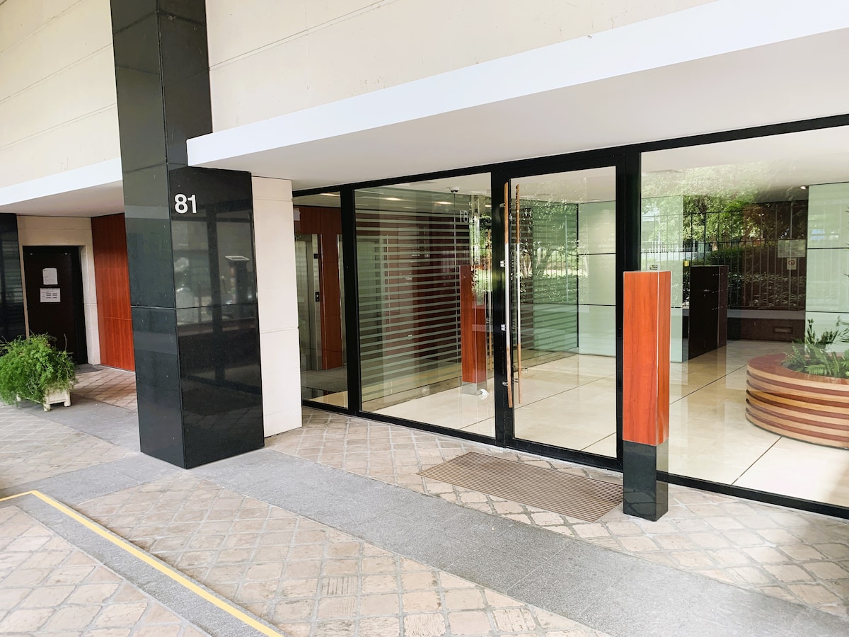 A modern entrance to the building is framed by sleek glass doors and elegant wooden accents. The polished stone flooring leads into a well-lit lobby area, featuring an elevator and greenery elements on either side, creating a welcoming entryway.