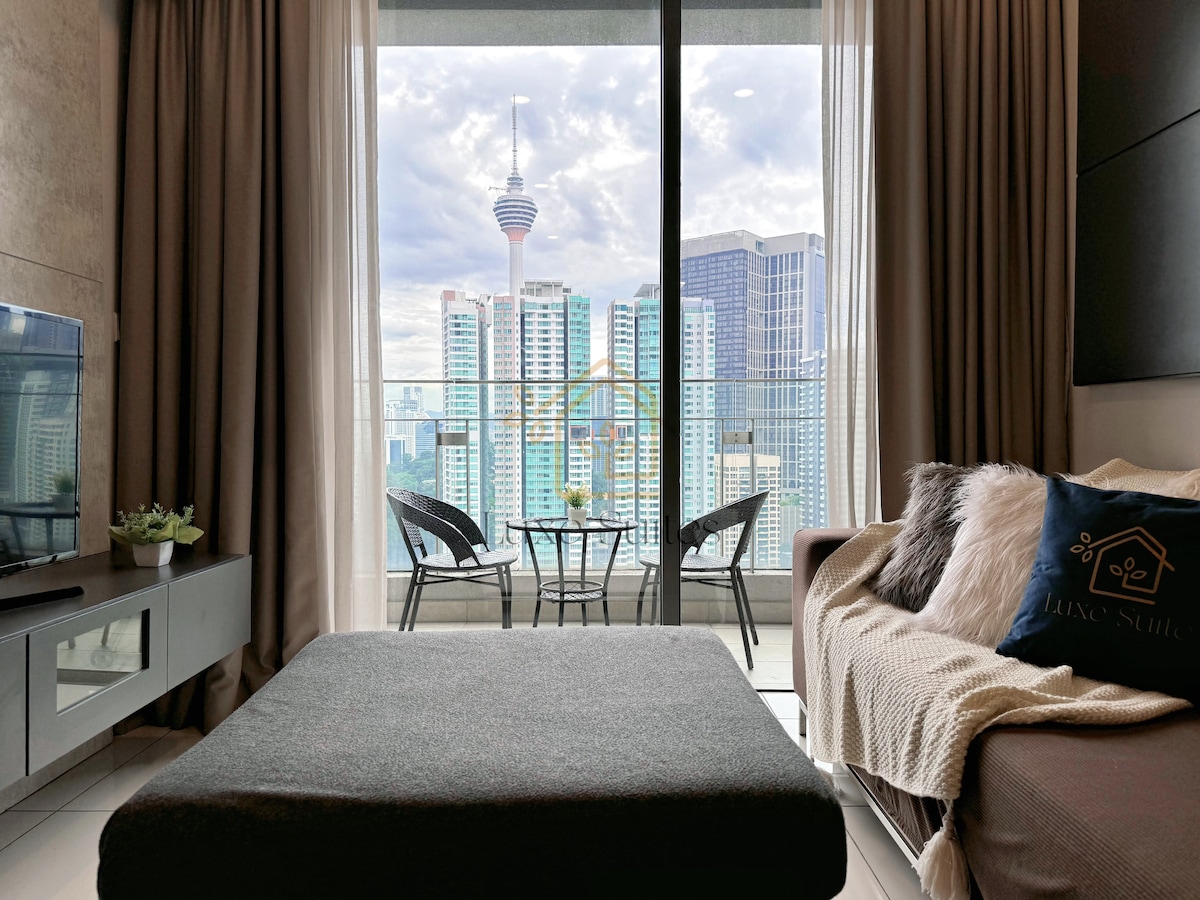 A cozy living area features a gray sofa positioned near a large window. Outside, city skyscrapers are visible along with the iconic Petronas Towers. A small balcony with two chairs and a table complements the view, creating a space for relaxation.