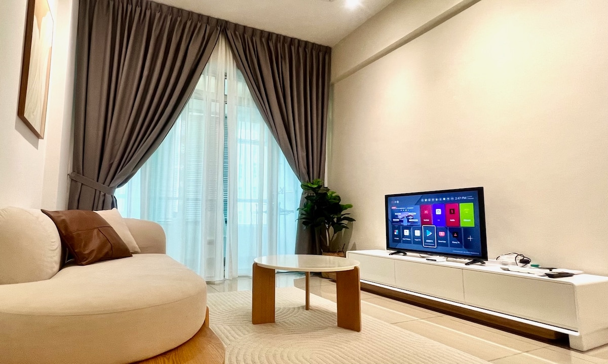 A living room area is defined by a spacious sofa accompanied by a round coffee table. A large LED TV is positioned on a white cabinet, with a greenery accent plant nearby. Floor-to-ceiling curtains frame the windows, allowing natural light to filter through.
