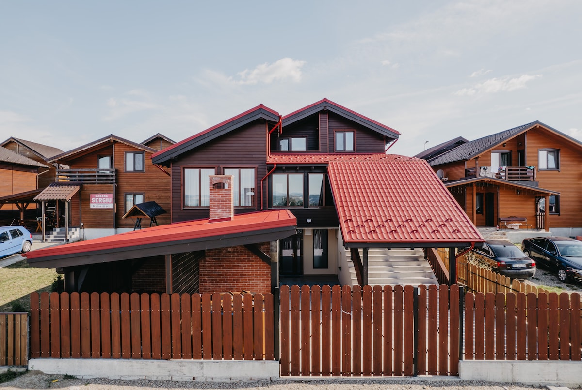 The exterior of the property showcases a modern structure with a red-tiled roof and large windows. A welcoming entrance is framed by a wooden fence, with parking areas visible in front of the building. Surrounding wooden cabins enhance the rustic charm of the setting.